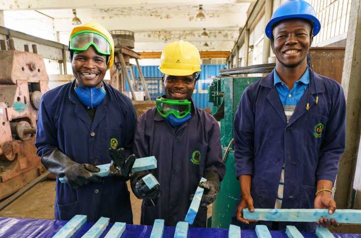 Three smiling young men wearing protective equipment in a warehouse hold columns of soap ready to be cut into bars 